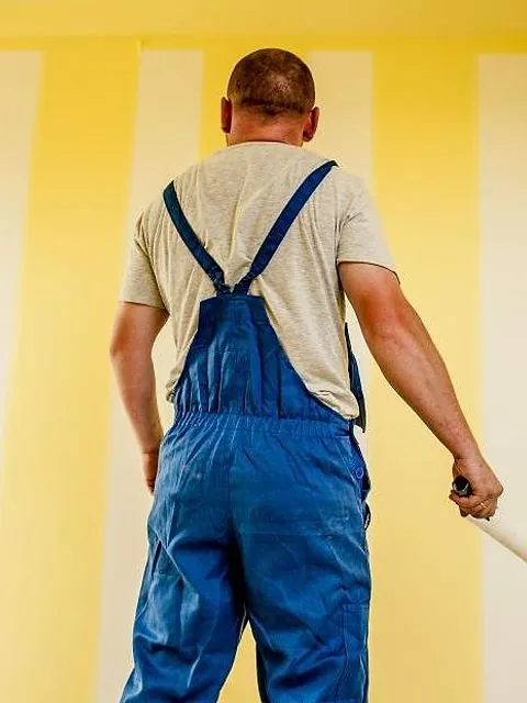 Peintre en salopette vu de dos devant un mur jaune