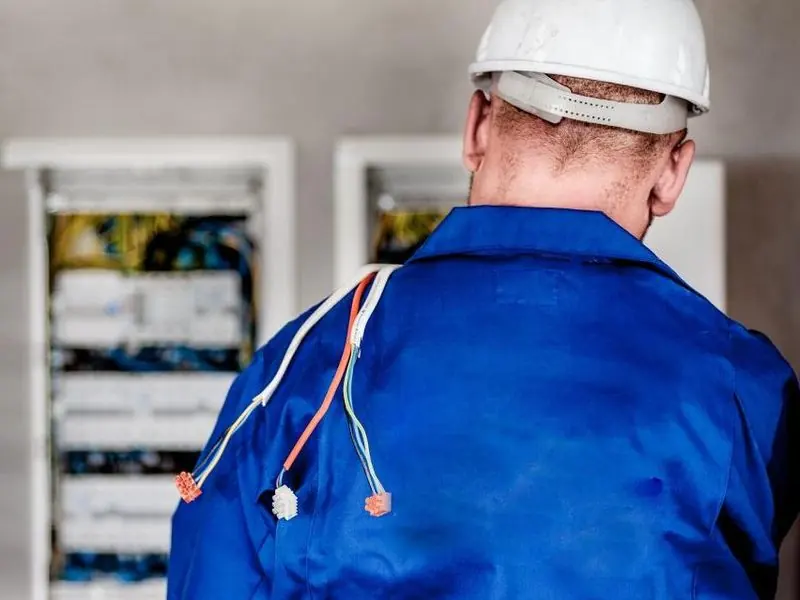 Électricien vu de dos, casque blanc, devant un tableau électrique ouvert, avec des fils sortant de sa veste