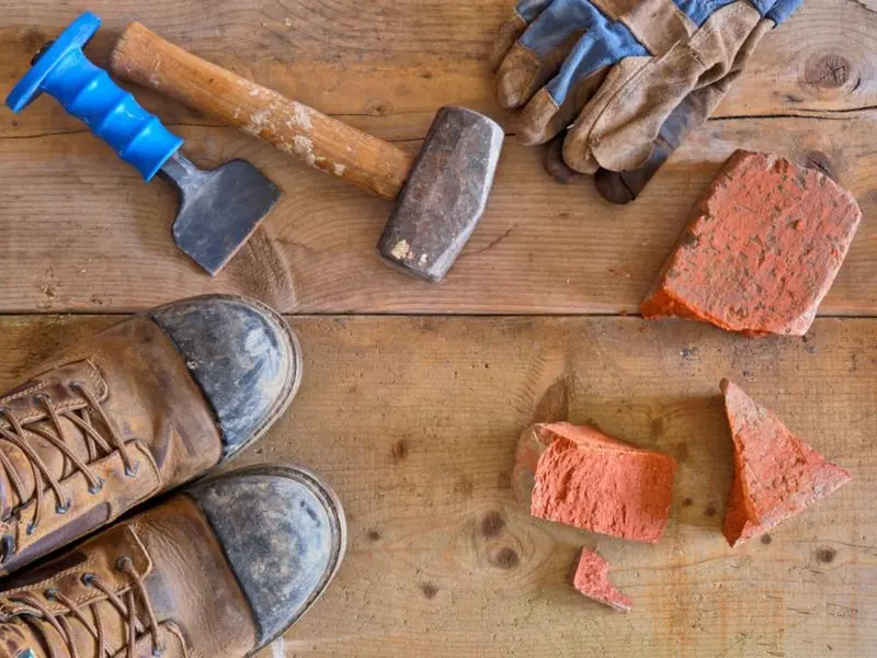 Outils de maçon posés sur du bois : burin, masse, gants de travail, chaussures de sécurité et morceaux de brique