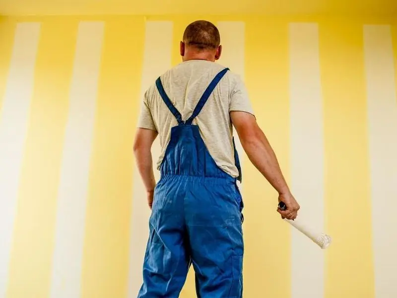 Peintre en salopette vu de dos devant un mur jaune à rayures blanches, un rouleau à la main